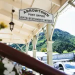 Arrowtown Post Office hanging sign under a covered walkway with mountains behind.