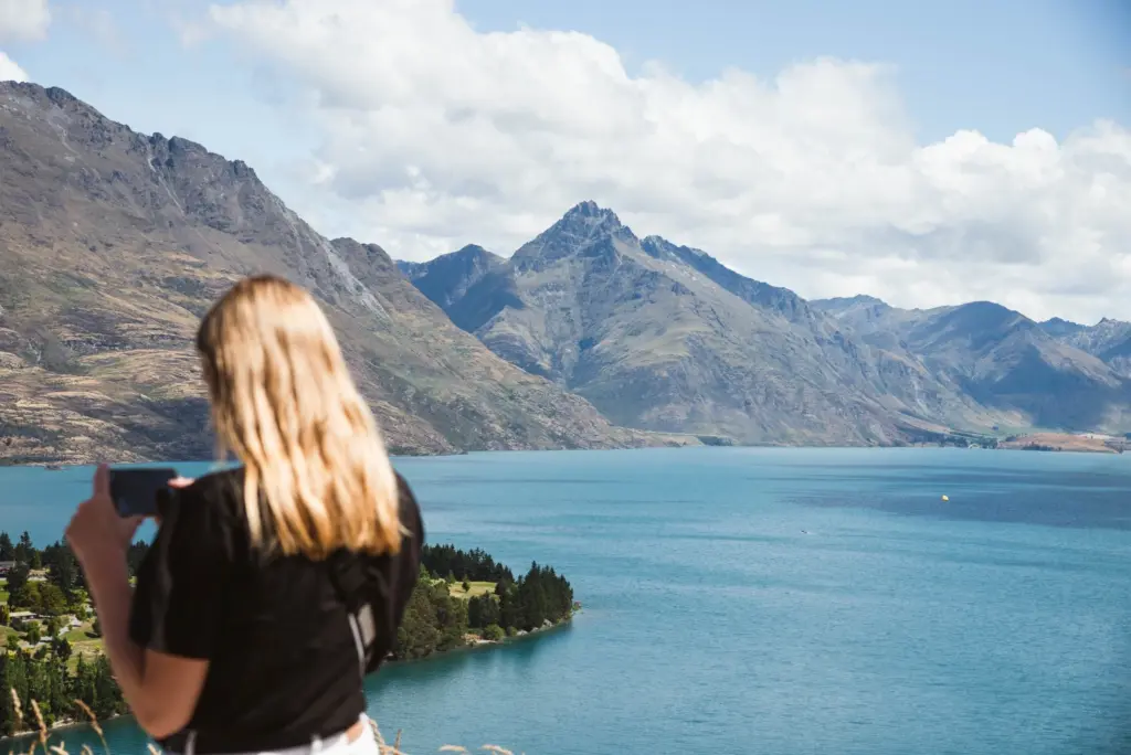 Traveller holding a phone while looking over Lake Wakatipu and surrounding mountains.