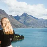 Traveller holding a phone while looking over Lake Wakatipu and surrounding mountains.