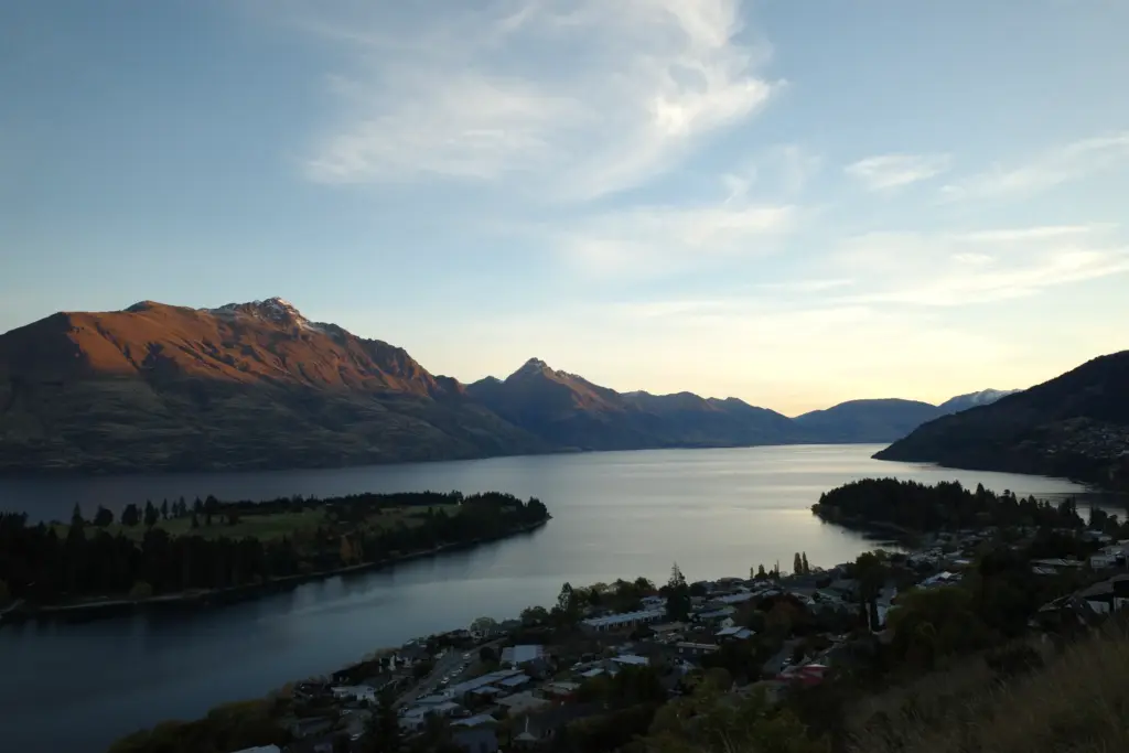 Evening panorama of Lake Wakatipu and Queenstown with mountain silhouettes and soft sunset light.