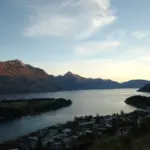 Evening panorama of Lake Wakatipu and Queenstown with mountain silhouettes and soft sunset light.